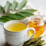 A warm cup of soursop leaf tea with dry leaves and a honey jar on white marble